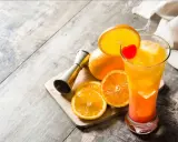 Kolorowy drink tequila sunrise z pomarańczą i wisienką, obok pomarańcze i miarka barmańska.