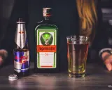 Jägermeister z czym pić? Klasycznie z Red Bullem, tworząc popularny "Jägerbomb". Na stole butelka Jägermeistera, Red Bull i szklanka z drinkiem.