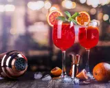 Dwa kieliszki orzeźwiającego drinka z prosecco z czymś czerwonym, plasterkami pomarańczy i miętą. Obok shaker i pomarańcze.