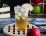 Orzeźwiający drink z sokiem jabłkowym, lodem i plasterkiem jabłka. W tle skrzynka z jabłkami.