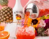 Malibu z sokiem ananasowym nalewany do szklanki z lodem. W tle ananas, butelka Malibu i kwiaty.