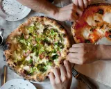 Dwie pizze na stole, jedna z brukselką i boczkiem, druga margherita. Ludzie sięgają po kawałki, pokazując, ile wyrasta ciasto na pizze.