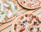 Trzy pizze na drewnianym stole. Jedna z szynką parmeńską i rukolą, druga z salami i pieczarkami, trzecia z pieczarkami. Szukasz alternatywy, jaki ser do pizzy zamiast mozzarelli?