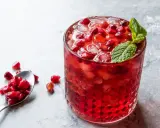 Orzeźwiający drink z grenadyną, lodem i pestkami granatu, ozdobiony listkiem mięty. Obok łyżeczka z owocem.