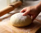 Dłoń dotyka kulki ciasta na pizzę, posypanej mąką. Obok wałek. Czy ciasto na pizzę można mrozić? Tak, można je zamrozić.
