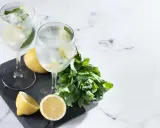 Dwa orzeźwiające drinki z prosecco, lodem, cytryną i miętą, prezentowane na czarnej desce z dodatkiem świeżych cytryn i gałązki mięty.