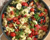 Kolorowa sałatka z tortellini, pomidorkami koktajlowymi, czarnymi oliwkami, papryką i natką pietruszki w dużej patelni.
