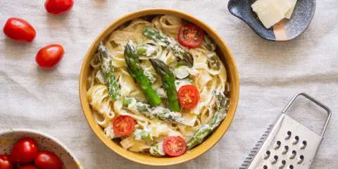 Pyszny makaron ze szparagami w kremowym sosie, ozdobiony pomidorkami koktajlowymi. Obok miseczka z parmezanem i tarką.