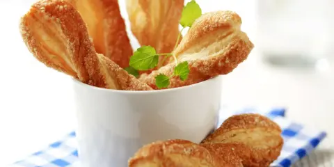 Chrupiące paluchy z ciasta francuskiego posypane cukrem, podane w białym naczyniu na niebiesko-białej kratce.