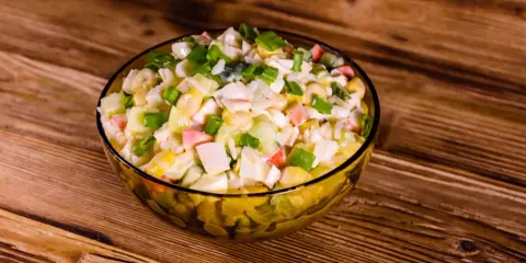 Pyszna sałatka z surimi, z kukurydzą, jajkiem i szczypiorkiem, podana w szklanej misce na drewnianym blacie.