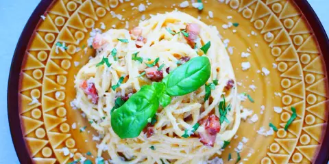 Pyszne spaghetti carbonara ze śmietaną, posypane parmezanem i natką pietruszki, ozdobione listkiem bazylii.