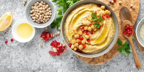 Pyszny hummus z ciecierzycy, udekorowany pestkami granatu i natką pietruszki, obok cytryny i oliwy.