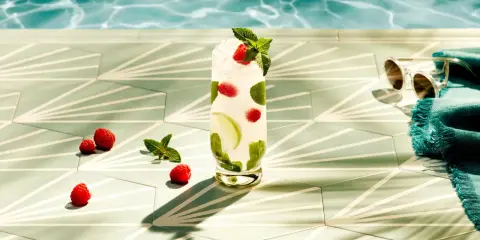 Orzeźwiający drink z malinami i miętą, idealny do Bacardi malinowego. Obok leżą maliny, a w tle widać basen i okulary przeciwsłoneczne.
