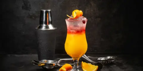 Kolorowy drink Sex on the Beach z pomarańczą i żurawiną, obok shakera i akcesoriów barmańskich. Idealny przepis na lato!