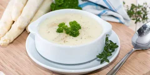 Aksamitny krem ze szparagów, udekorowany natką pietruszki, obok świeżych szparagów i łyżki.