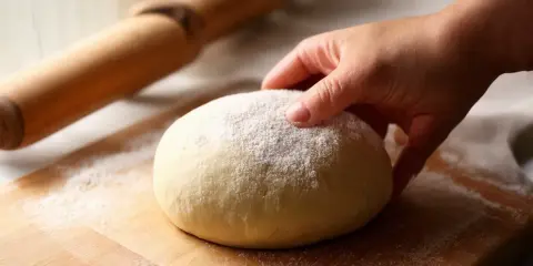 Dłoń dotyka kulki ciasta na pizzę, posypanej mąką. Obok wałek. Czy ciasto na pizzę można mrozić? Tak, można je zamrozić.
