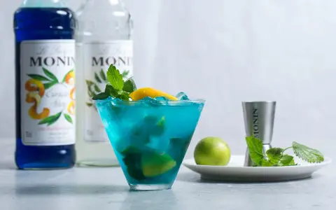 Orzeźwiający drink z syropem blue curacao smak, ozdobiony miętą i cytryną. Obok butelki syropu Monin.