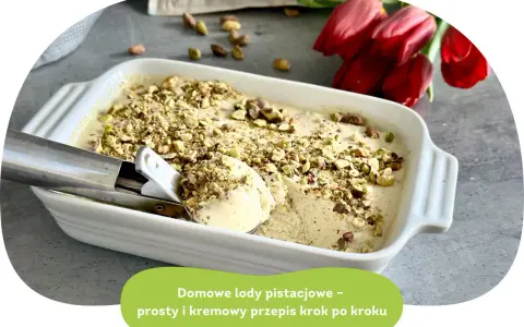 Kremowe lody pistacjowe w białej formie, posypane orzechami. Łyżka właśnie nabiera porcję tego pysznego deseru.