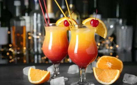 Dwa kolorowe, domowe drinki z wódką, ozdobione pomarańczą i wiśnią, z kostkami lodu i owocami w tle.