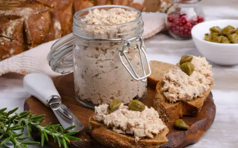 Słoik pełen pysznej pasty z tuńczyka, gotowej do rozsmarowania na chlebie. Na desce kromki z pastą i kaparami, obok gałązka rozmarynu.