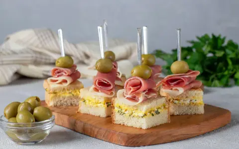 Eleganckie mini przekąski na imprezę: kanapki z szynką, jajkiem i oliwkami na drewnianej desce.