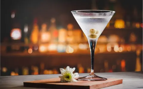 Eleganckie drinki z martini, ozdobione oliwkami, stoją na blacie baru. Obok kieliszka leży biały kwiat.