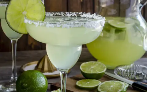 Orzeźwiająca margarita z solą na brzegu kieliszka, ozdobiona plasterkiem limonki. Obok dzbanek z drinkiem i świeże limonki.