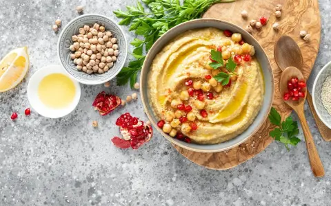 Pyszny hummus z ciecierzycy, udekorowany pestkami granatu i natką pietruszki, obok cytryny i oliwy.