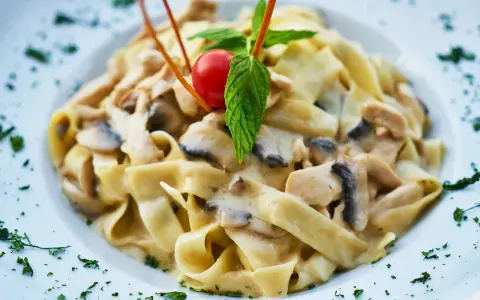 Pyszny makaron tagliatelle z kremowym sosem pieczarkowym, udekorowany pomidorkiem koktajlowym i listkiem mięty.