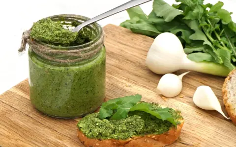 Słoik domowego pesto z rukoli, łyżka w środku, kromka chleba z pastą i świeże składniki: czosnek i rukola.
