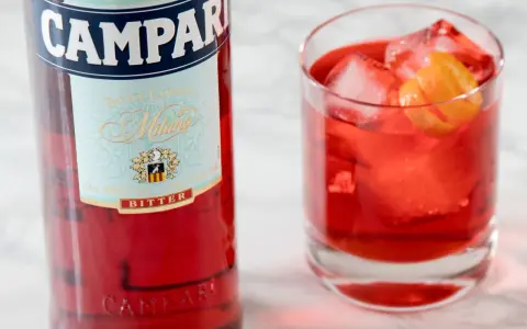 Butelka Campari i szklanka z czerwonym drinkiem, lodem i skórką pomarańczy. Idealny aperitif, który może być bazą do drinków z Aperolem.