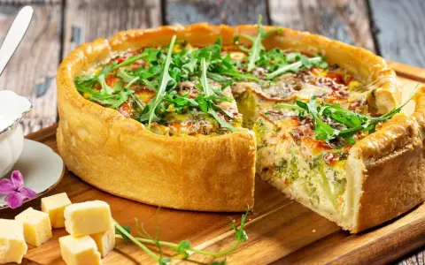 Pyszna tarta z brokułami i łososiem, udekorowana rukolą. Obok kawałki sera i sos.