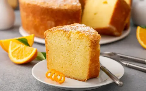 Puszysta babka pomarańczowa, oprószona cukrem pudrem, z plasterkami pomarańczy i ozdobną skórką na talerzyku.