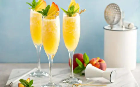 Trzy orzeźwiające drinki mimoza w kieliszkach flute, ozdobione brzoskwinią i miętą, na marmurowym blacie.