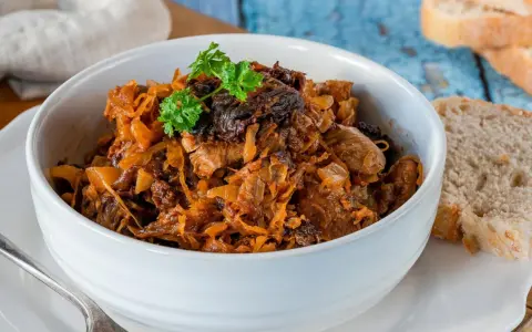 Pyszny bigos w białej misce, z gałązką natki pietruszki. Obok kromki chleba. Idealny na zimowe wieczory.
