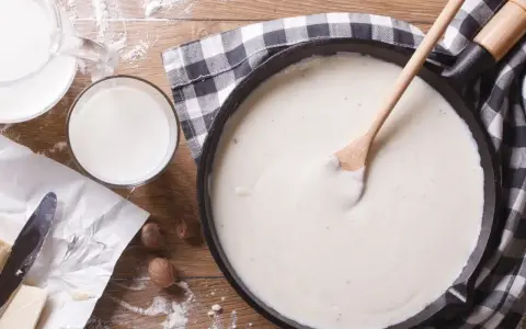 Gładki sos beszamelowy w patelni, obok mleko, masło i gałka muszkatołowa. Idealny do zapiekanek.