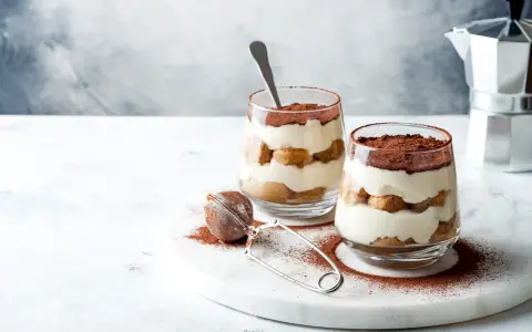 Dwa pucharki z tiramisu bez jajek, posypane kakao, na marmurowej desce.