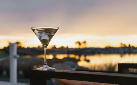 Idealny martini wstrząśnięte nie mieszane, z oliwką, na tle zachodzącego słońca nad wodą.