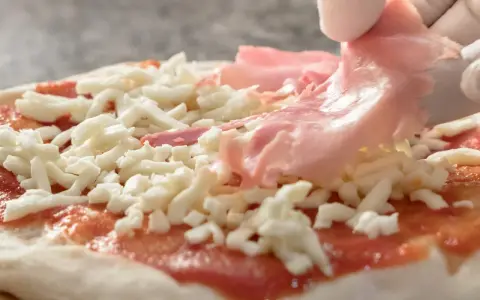 Ręce w białych rękawiczkach układają cienkie plastry szynki do pizzy na serze i sosie pomidorowym.