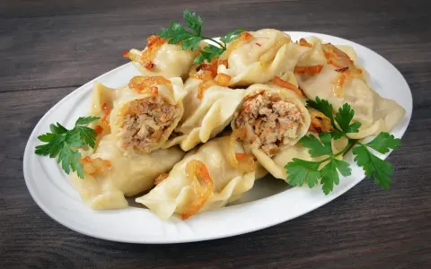 Pyszne pierogi z mięsem i kapustą, okraszone skwarkami i udekorowane natką pietruszki, na białym talerzu.