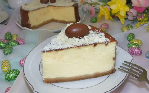 Pyszny sernik z mascarpone, udekorowany czekoladowymi jajeczkami i wiórkami białej czekolady, na tle wielkanocnych ozdób.
