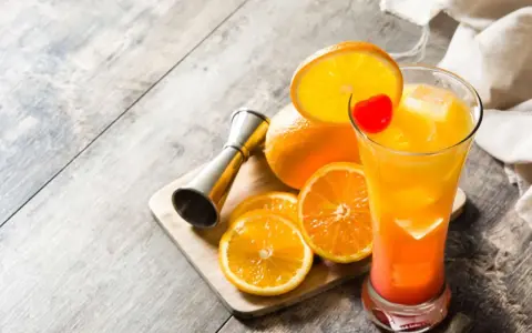 Kolorowy drink tequila sunrise z pomarańczą i wisienką, obok pomarańcze i miarka barmańska.