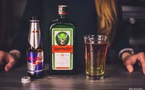 Jägermeister z czym pić? Klasycznie z Red Bullem, tworząc popularny "Jägerbomb". Na stole butelka Jägermeistera, Red Bull i szklanka z drinkiem.