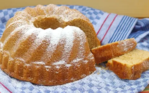 Puszysta babka wielkanocna, oprószona cukrem pudrem, leży na niebiesko-białym ręczniku. Obok niej kilka kromek ciasta.