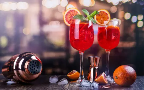 Dwa kieliszki orzeźwiającego drinka z prosecco z czymś czerwonym, plasterkami pomarańczy i miętą. Obok shaker i pomarańcze.