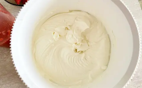 Gładki, puszysty krem z mascarpone w białej misie, gotowy do deseru. Obok miski widać miseczkę z truskawkami.