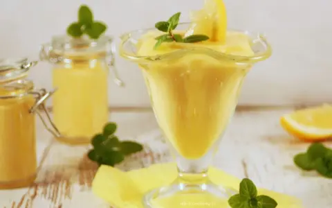 Aksamitny lemon curd w pucharku, ozdobiony plasterkiem cytryny i miętą. Obok słoiczki z tym samym kremowym przysmakiem.