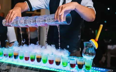 Barman nalewa kolorowe shoty z wielopoziomowego shakera. Efektowny drink bar diy z dymem i neonowym oświetleniem.