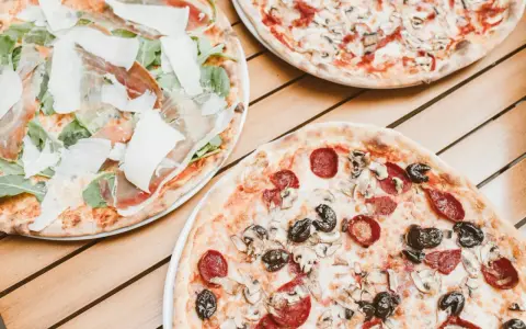 Trzy pizze na drewnianym stole. Jedna z szynką parmeńską i rukolą, druga z salami i pieczarkami, trzecia z pieczarkami. Szukasz alternatywy, jaki ser do pizzy zamiast mozzarelli?