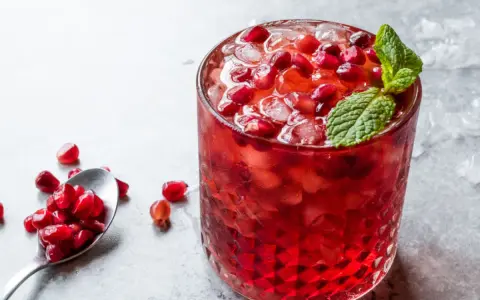 Orzeźwiający drink z grenadyną, lodem i pestkami granatu, ozdobiony listkiem mięty. Obok łyżeczka z owocem.